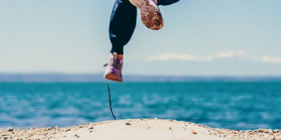 person in black pants and brown shoes jumping on brown rock near body of water during